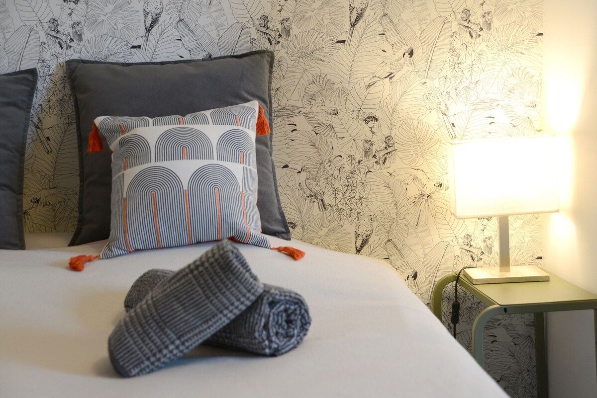 Détail du lit : linge blanc, coussin décoratif à pompons orange et serviette roulée sur papier peint jungle — La Maison des Coccinelles