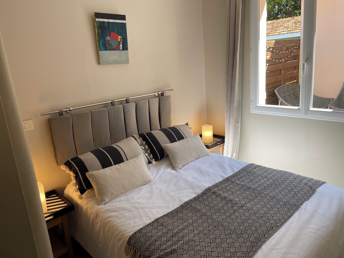 Chambre avec lit double, tête de lit grise capitonnée et fenêtre donnant sur la terrasse extérieure — La Maison des Coccinelles