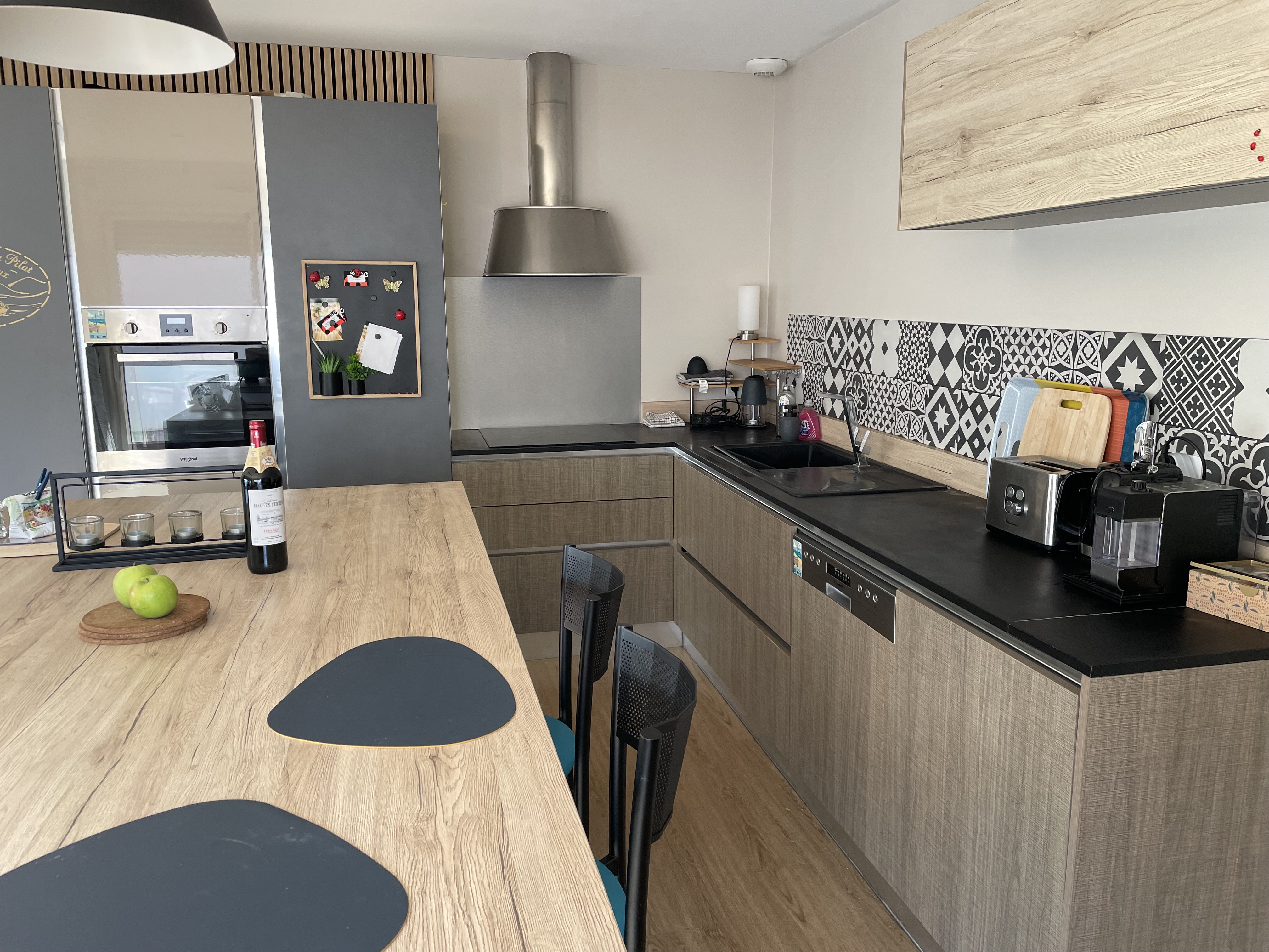 Cuisine équipée avec meubles en bois clair, plan de travail noir, hotte inox et crédence carreaux de ciment — La Maison des Coccinelles