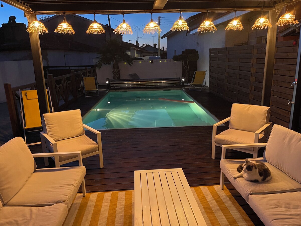 Terrasse piscine au crépuscule avec guirlandes lumineuses allumées, canapé et fauteuils blancs — La Maison des Coccinelles