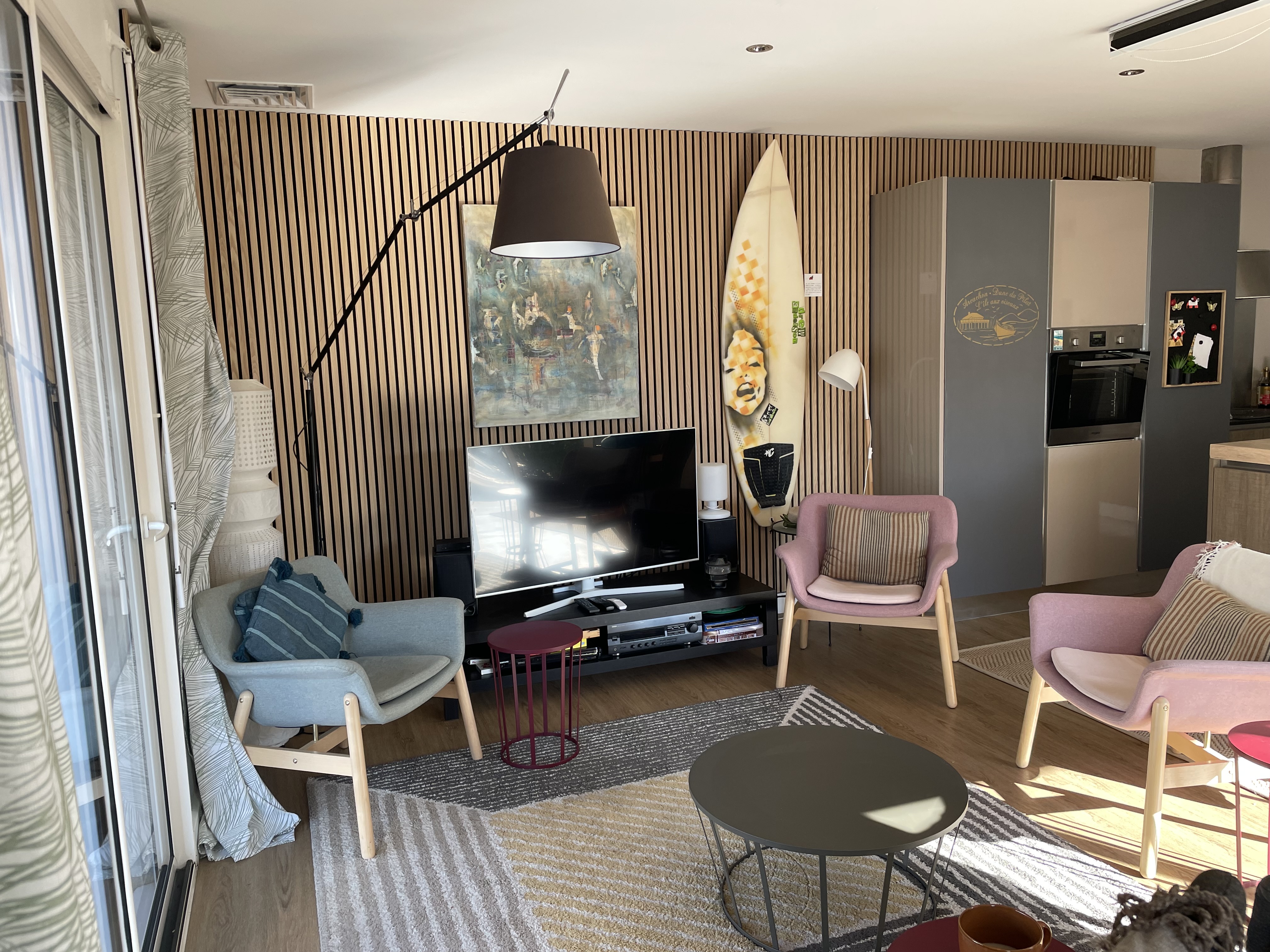 Salon lumineux avec télévision, planche de surf décorative, fauteuils gris et rose sur mur à tasseaux en bois — La Maison des Coccinelles