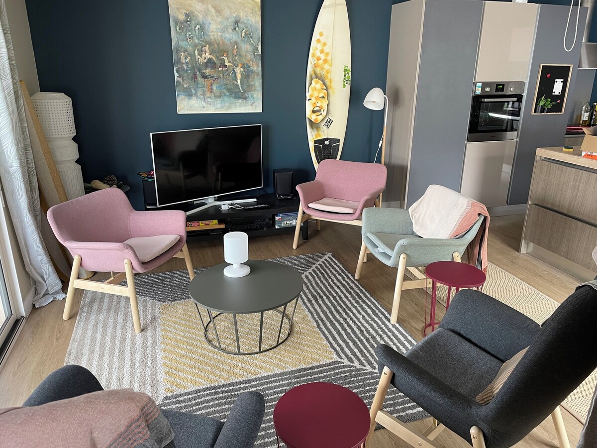 Salon avec fauteuils roses, télévision, table basse ronde et décoration planche de surf — La Maison des Coccinelles